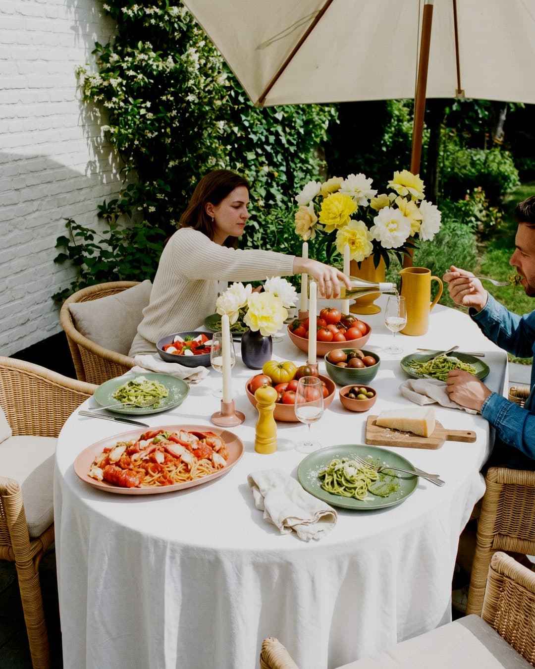 Sunlit beach tablescape on white linen with whole grilled fish, potatoes, tomatoes and guests sharing a meal