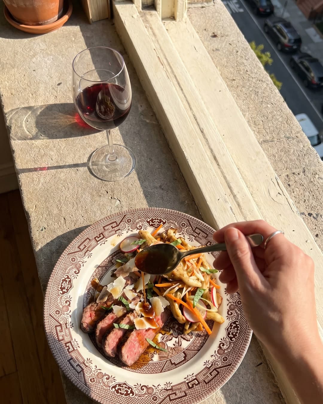A candid overhead snapshot of a patterned brown-and-cream ceramic plate resting on a sunlit stone windowsill, with a glass of red wine and a hand spooning sauce over sliced beef and mushrooms.
